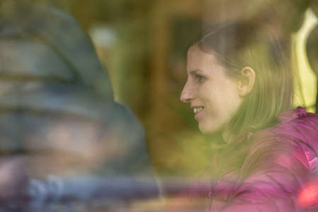 Profile view of a smiling young woman sitting inside a coffee shop talking to a friend. View through a window.の写真素材