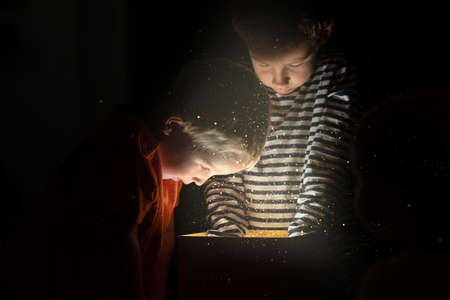 Two toddler boys standing over golden gift box with magical lights and glitters coming out reaching...