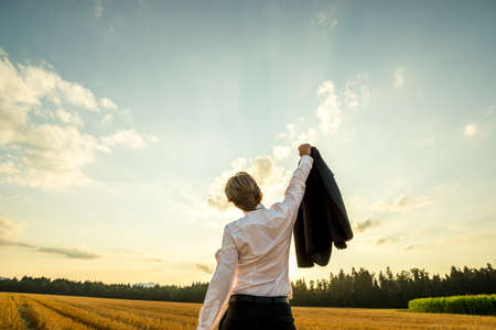 View from behind of a businessman standing in sawn field under evening sky with his arm holding suit jacket raised up high in triumph or celebration.の写真素材