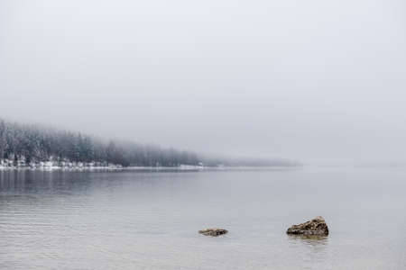 Two rocks peeking out calm winter lake with forest in background on a foggy day.の写真素材