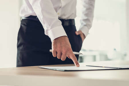 Entrepreneur standing at his office desk holding an ink pen pointing to a document or subscription form to a signature line.の写真素材