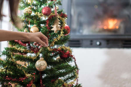 Female hand decorating Christmas tree by the warm fire in living room fireplace.