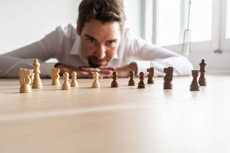Businessman leaning his head on his palms on office desk looking at white against black chess pieces arranged on wooden desk with copy space.の写真素材