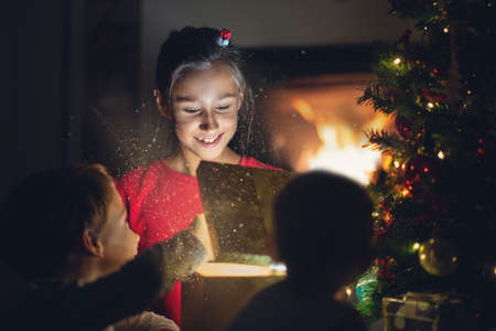 Little girl joyfully smiling as she opens golden gift box under christmas tree with her siblings...