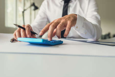 Architect or designer sitting at his office desk using calculator as he works on his project holding a pencil and ruler on his table.の写真素材
