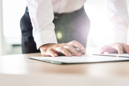 Businessman standing at his office desk leaning in to sign a document holding an ink pen with lens flare and bright background.の写真素材