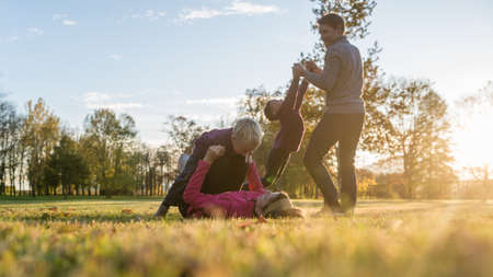 Young family of four playing in autumn park with mother lying in grass lifting her toddler son and father spinning the other son around holding his hands.の写真素材