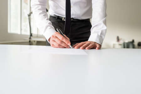 Front view of businessman standing behind his office desk signing a document or contract.の写真素材