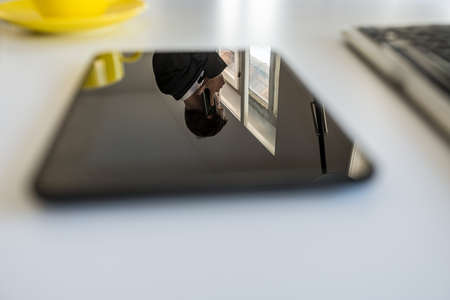 Reflection of a young businessman talking on a telephone in a black digital tablet lying on an office desk.の写真素材