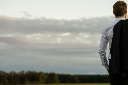 Young businessman holding his suit jacket over his shoulder  standing at the left side of the image outside in nature looking into the distance.の写真素材