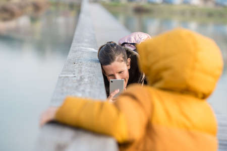 Young mother taking a photo of her toddler child outside by a wooden fence with her mobile phone.の写真素材