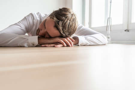 Tired businessman resting his head on his hands sitting at wooden office desk. With copy space on...