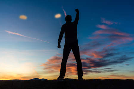 View from behind of a man standing on top of a wall with his arm raised high in triumph under a...