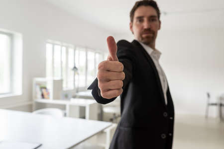 Young businessman standing in his bright office making a thumbs up gesture toward the camera.の写真素材