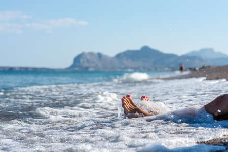 Closeup of female feet with pink pedicure in a foamy sea of a wave on pebble beach of the island Rhodes.の写真素材