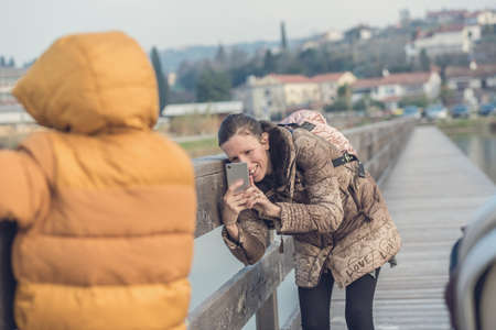 Happy young mother taking a photo with her mobile phone of her toddler child outside on a wooden path by the sea.の写真素材