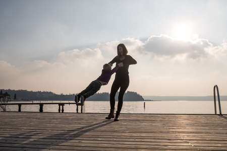 Happy young mother standing on wooden pier holding her toddler son by his hands spinning him around...