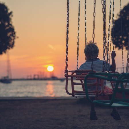 Toddler boy riding a carousel with red sun setting in to a sea on a summer evening.