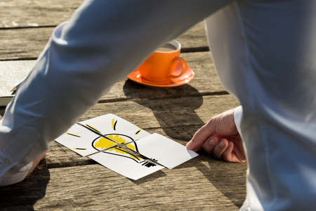 View through a leaning arm of a businessman assembling a light bulb drawn on four paper pieces on wooden desk.の写真素材