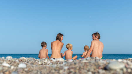 Young family of five sitting on a pebble beach in beautiful Greece with their backs to the camera enjoying their holidays.の写真素材