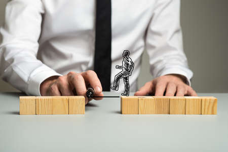 Business executive drawing a line between two separated piles of wooden blocks for a silhouetted businessman to walk across in a conceptual image.の写真素材