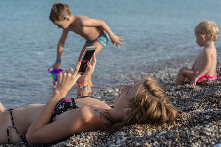 Young mother lying on pebble beach using her mobile phone uninterested in her two kids as they play around her.の写真素材