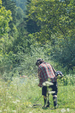 View from behind of a man in beautiful green nature mowing high grass wearing protection for the...