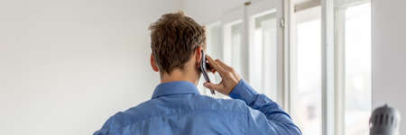 View from behind of young businessman standing by the office windows talkin on mobile phone.