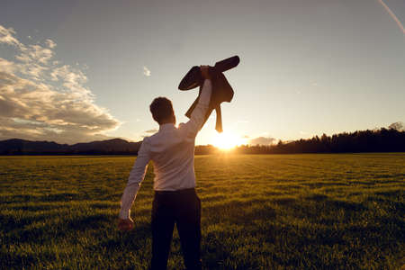 View from behind of a young businessman standing in beautiful field lit by the setting sun raising his suit jacket high up in the air in triumph.の写真素材