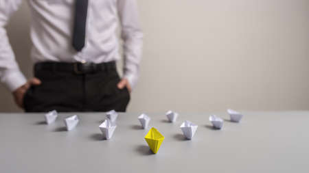 Business leader standing next to a desk with a group of origami paper boats, yellow one leading the others.の写真素材