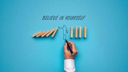 Believe in yourself sign over a hand drawn man stopping falling dominos in a conceptual image. Over blue background.の写真素材