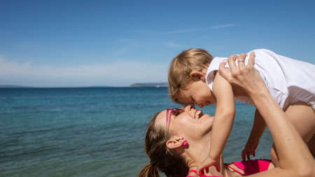Happy young mother playing with her toddler daughter in diapers lifting her by the spring sea.の写真素材