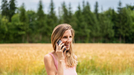 Young woman with a smile on her face talking on mobile phone as she takes a walk in nature. With wheat field in background.の写真素材