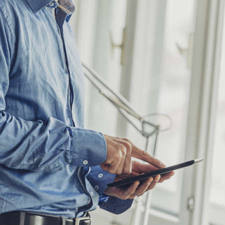 Businessman standing by office windows browsing on his digital tablet.