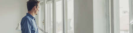 Wide view image of young businessman standing by bright office windows looking out.