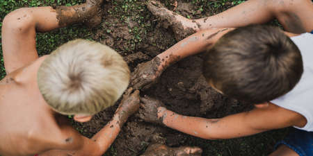 Top view of two toddler boys digging a hole with their hands in a muddy ground.