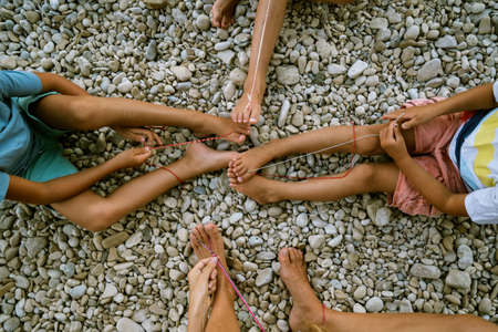 Top view of four children sitting on pebbles making knotted bracelets of colorful strings sticked around their toes.の写真素材
