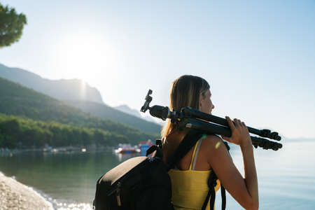 Young female photographer standing by the sea in the early morning with photographic backpack and...