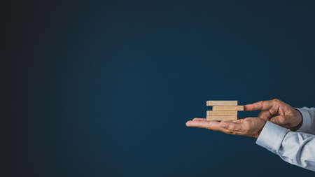 Businessman stacking wooden pegs in the palm of his hand. Over navy blue background with copy space.の写真素材