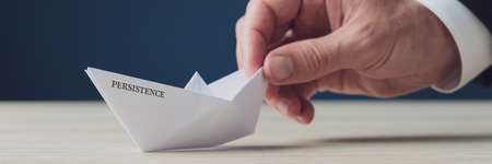 Wide view image of businessman hand holding paper made origami boat with Persistence sign on it in a...