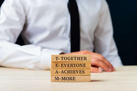 Team - together everyone achieves more sign on stacked wooden pegs wit businessman sitting in...