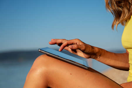 Closeup side view of a woman sitting on the beach browsing on her digital tablet on a bright sunny day.の写真素材