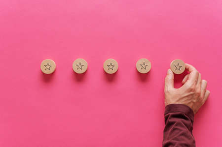 Male hand placing five wooden cut circles with star shape on them in a row in a conceptual image of quality and satisfaction. Over pink background.の写真素材