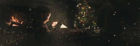 Wide view image of an excited  boy opening a holiday gift box under Christmas tree next to a fireplace. With glitters and fairy dust all over the image.の写真素材