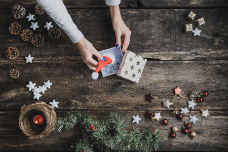 Top view of female hand placing red Santa hat and silver stars in a gift box on a rustic wooden desk with holiday decoration, candle and pine tree branches on it.の写真素材