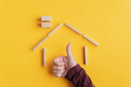 Male hand making a thumbs up gesture holding his hand inside a house made of wooden pegs and blocks. Over yellow background with copy space.の写真素材