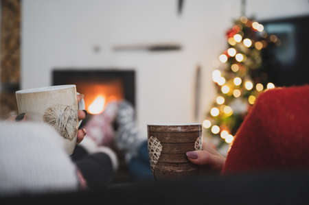 View from behind of two girlfriends sitting with their feet up by the fireplace next to glowing holiday tree drinking tea or coffee.の写真素材