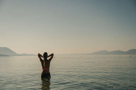View from behind of a young woman standing in peaceful morning sea going through her long hair with her fingers. の写真素材
