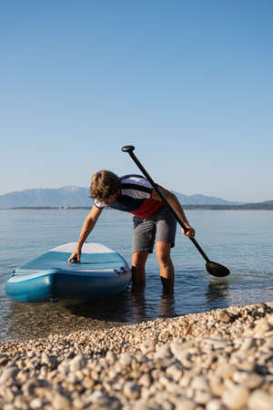 Low angle view of young man taking his sup board out of the calm water of morning sea after...