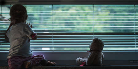 Toddler girl looking out the window through lowered venetian blinds with her teddy bear sitting next to her in a conceptual image.の写真素材